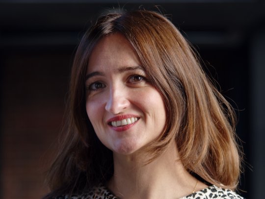 Dr. Denise Dekker Dr. Denise Dekker: eine Forscherin mit langen braunen Haaren, die eine schwarz-weiß gemusterte Bluse trägt. Sie steht vor einem dunklen Hintergrund.