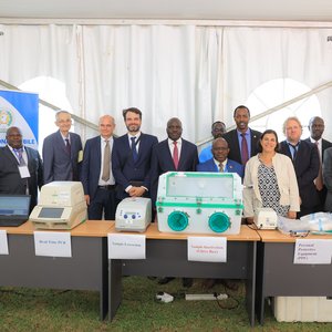 Eröffnung UVRI Exzellenzzentrum Auf dem Foto ist ein Tisch zusehen, auf dem Geräte wie eine PCR-Maschine und eine Glove Box stehen. Dahinter stehen 12 gut gekleidete Personen. Flankiert werden sie von zwei Postern des EAC Mobile Lab Projektes.