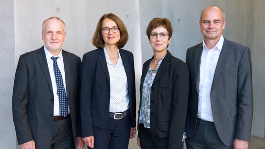 Gruppenfoto des BNITM-Vorstands mit Stephan Günther, Birgit Müller, Iris Bruchhaus, Jürgen May Foto der vier Vorstandsmitglieder: Zwei Frauen und zwei Männer stehen in dunklen Kostümen bzw. Anzügen gekleidet vor einer Betonwand und lächeln Richtung Kamera.