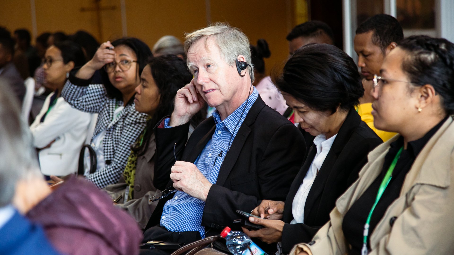 A man wearing a headset seated among other attendees during a CCSM 2024 conference session, appearing attentive, with others listening or looking at documents.