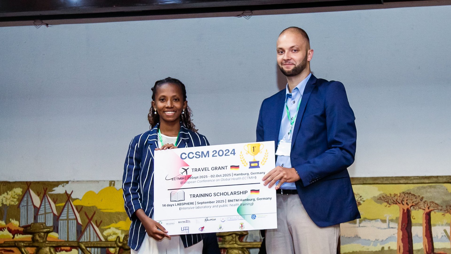 Two individuals standing together holding a large award certificate for the CCSM 2024 travel grant and training scholarship, with a background featuring decorative artwork.