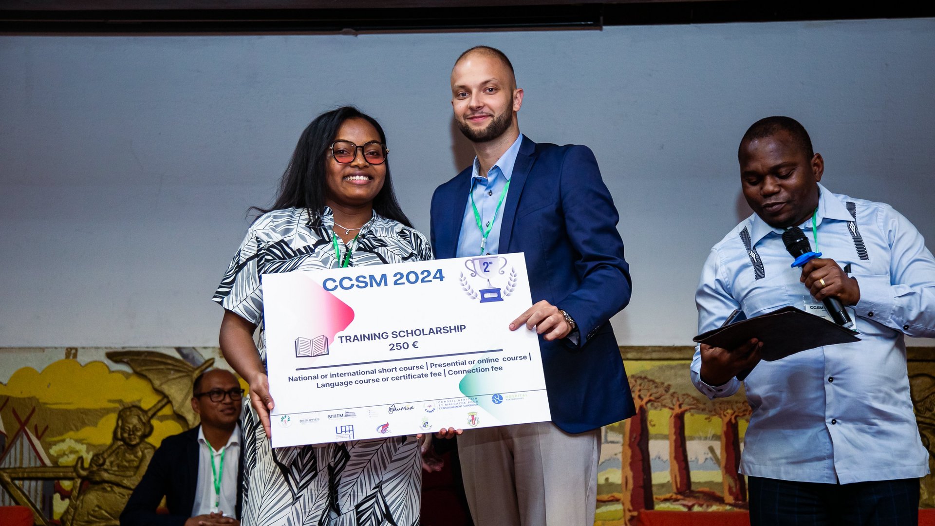 Two individuals posing with a large certificate for a CCSM 2024 training scholarship, while another person speaks into a microphone nearby on stage.