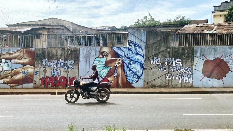 One road in Cotonou, capital of Benin A male Beninese rides a motorbike. Graffiti on the wall behind him says “wear a mask” and “Do not shake hands” in French. Next to that you see illustrations of hands using sanitizer, a female Beninese wearing a mask and a handshake with a red cross over it.