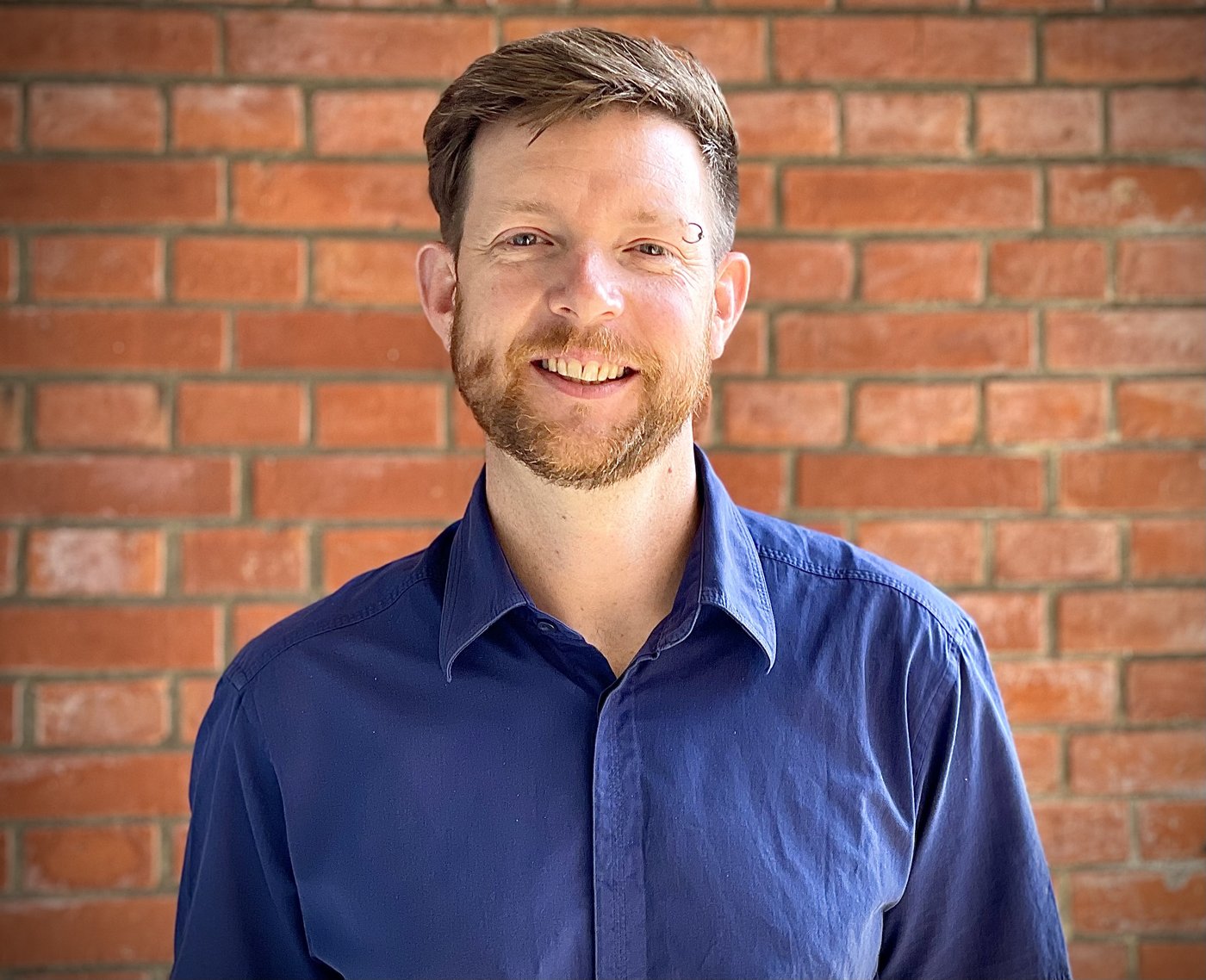 Dr. Benno Kreuels Benno Kreuels: a doctor with short reddish hair and a short reddish beard, wearing a dark blue casual shirt, stands in front of a brick wall. He wears a piercing ring above his left eyebrow.