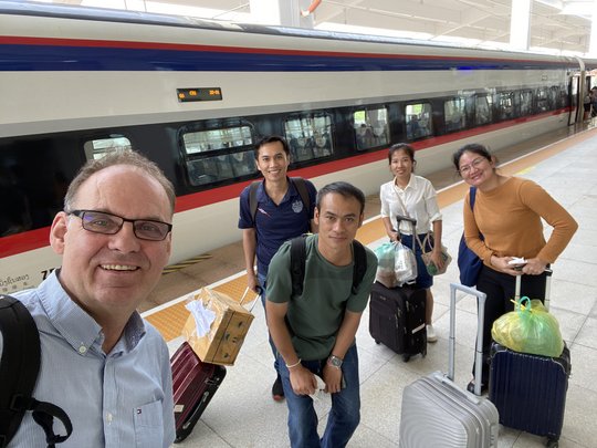The team before the train journey between South and North Laos The picture shows five people standing in front of a train with luggage and looking friendly into the camera.