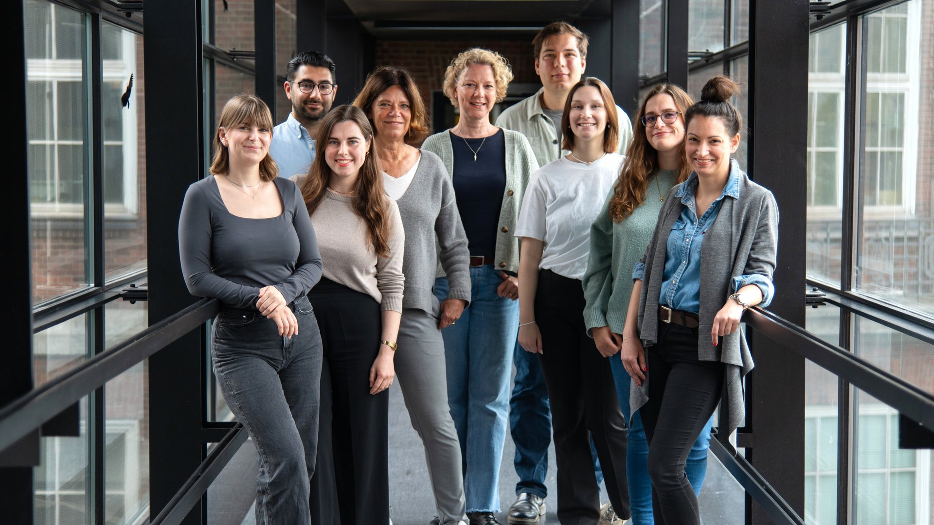 Molekulare Infektionsimmunologie 2025 In the picture, the 9 members of the working group are standing in the glass corridor from the old building to the new building of the Bernhard Nocht Institute for Tropical Medicine and smiling into the camera.