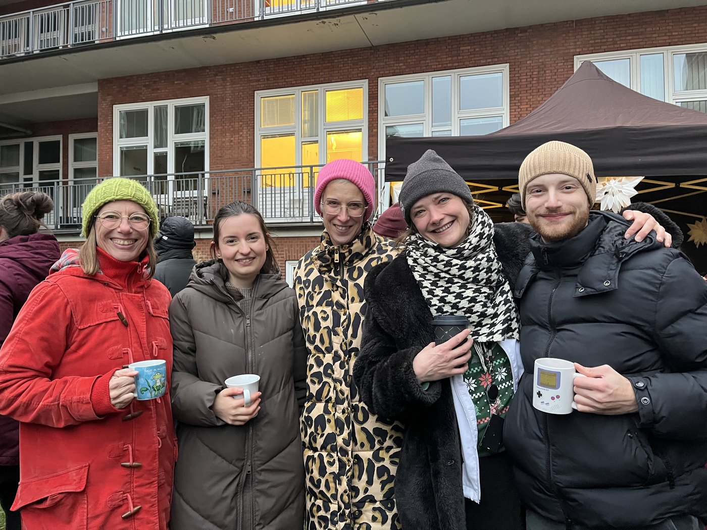 Five people in winter jackets with drinks in their hand in front of a red brick building