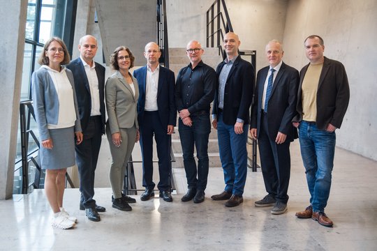 Erweteter Vorstand Eine Gruppe von acht Personen, fünf Männer und drei Frauen, steht in einer Reihe in einem Gebäude vor einer Betonwand. Sie tragen Business- und Freizeitkleidung und strahlen eine professionelle und zugleich entspannte Atmosphäre aus.