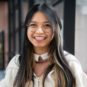 Bild von Mia Le - Sie trägt eine naturfarbene Jacke über einem roséfarbenen Pullover mit weißem Kragen. Sie hat dunkle, lange Haare, eine Brille und steht vor einer Fensterfront.