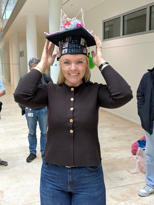 Lara Buer with doctoral hat The photo shows a close-up of Lara Buer after her exam. She is proudly holding her doctoral hat with both hands and smiling at the camera. The black cap, adorned with many colorful details, was handmade by her colleagues in Borstel. Lara is wearing a chic brown cardigan with gold buttons.