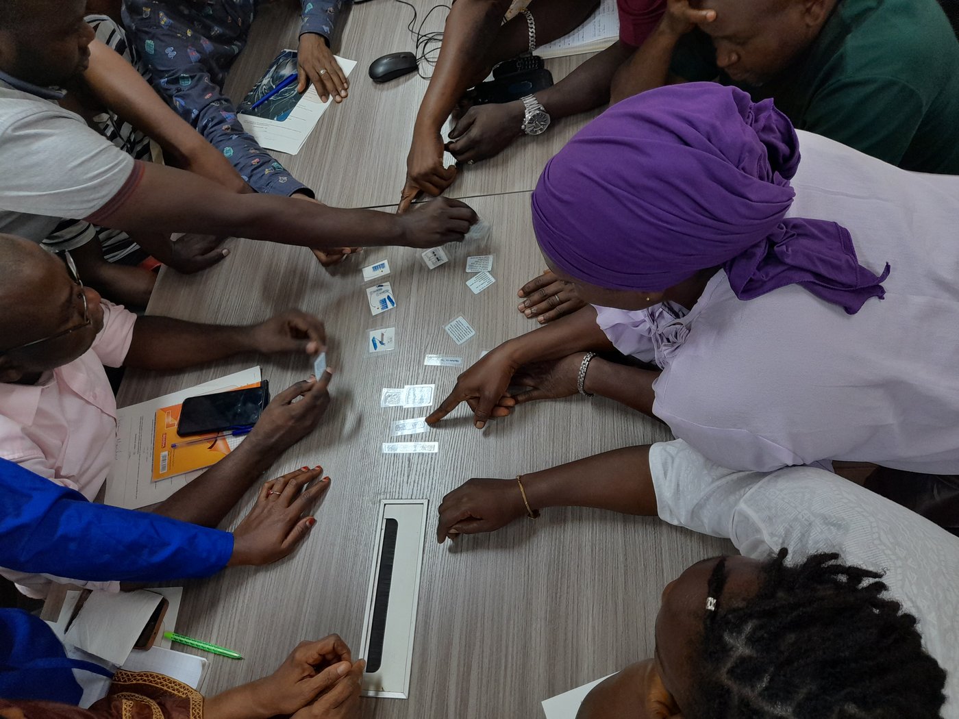 Ten Guinean lab staff in casual clothes are leaning over a table pointing at small plastic cards for an excercise. One person on the right wears a purple headscarf and a white shirt.
