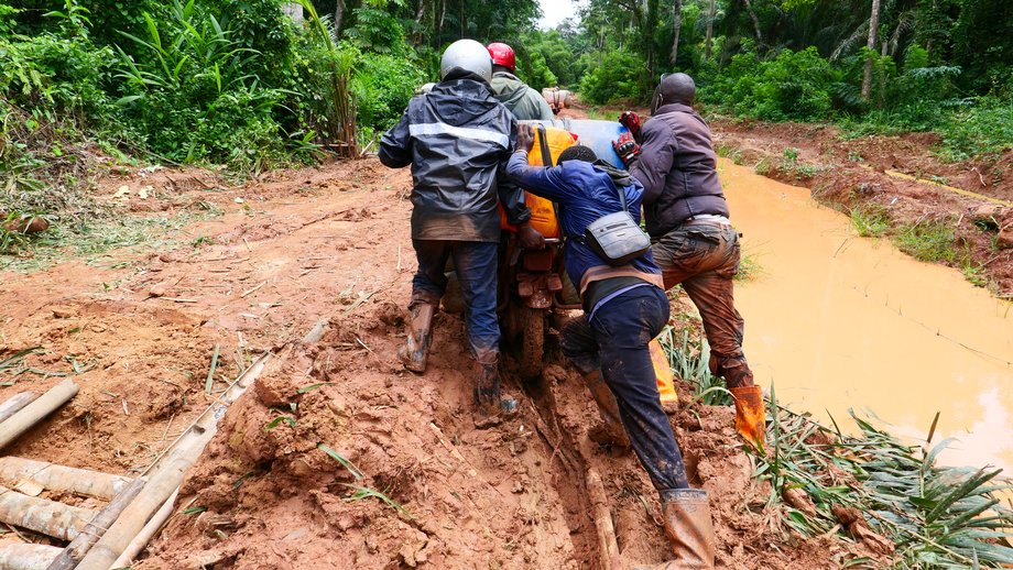 Reisen mit Motorrad in DRC Auf dem Foto sind vier Personen von hinten zu sehen, wie sie ein feststeckendes Motorrad durch Schlamm schieben.