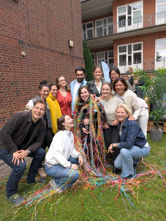 Group photo of AG Lotter and friends, summer party 2025 Barbara Honecker stands in the middle of her work group in the garden of the BNITM and is decorated with colourful streamers. Everyone in the picture is laughing happily into the camera