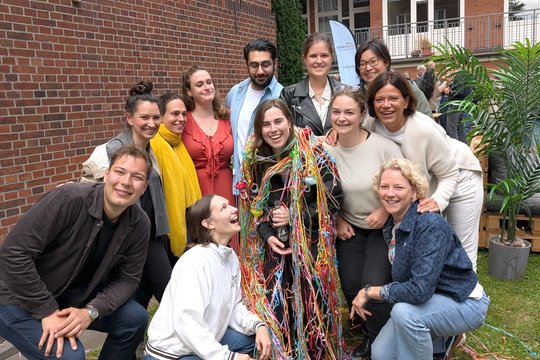 Group photo of AG Lotter and friends, summer party 2025 Barbara Honecker stands in the middle of her work group in the garden of the BNITM and is decorated with colourful streamers. Everyone in the picture is laughing happily into the camera