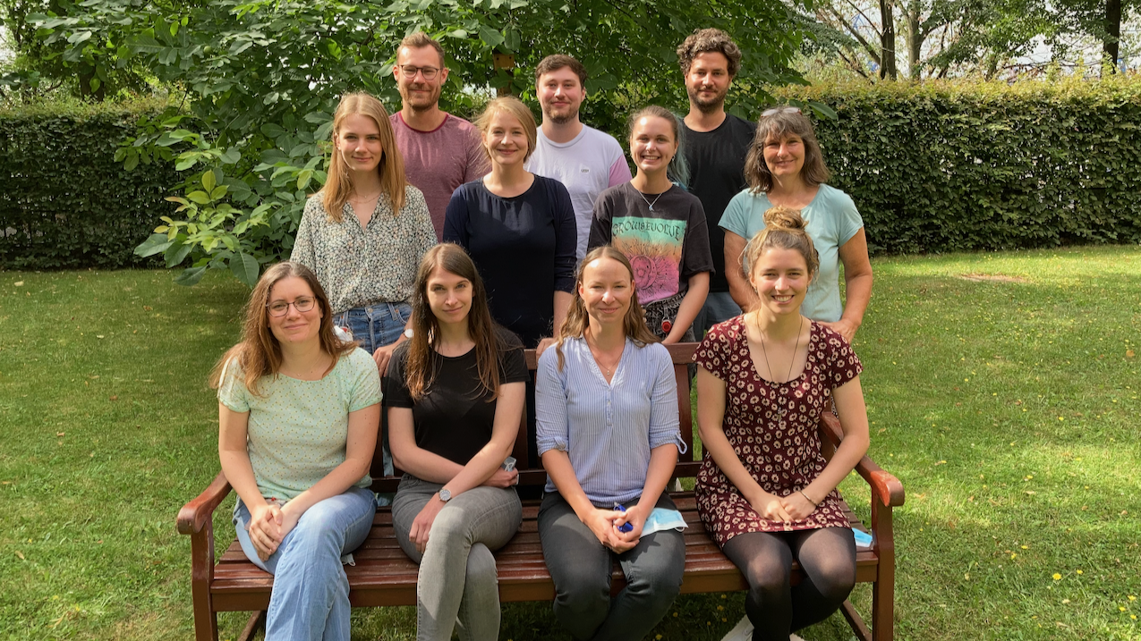 Sitzendes freundliches direktes Gruppenfoto im grünen Garten des BNITM. Die Personen sitzen teilweise auf eienr Parkbank andere stehen dahinter. Um sie herum ist Natur