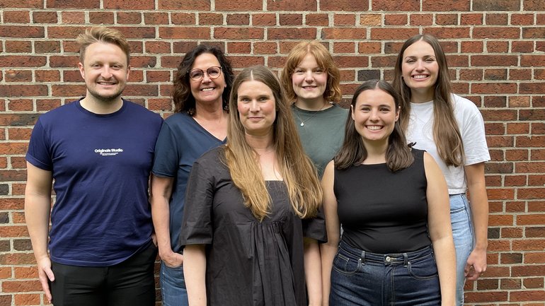 Laborgruppe Bachmann A group of six people smiles in front of a wall. They appear relaxed and happy and create a warm and friendly atmosphere.