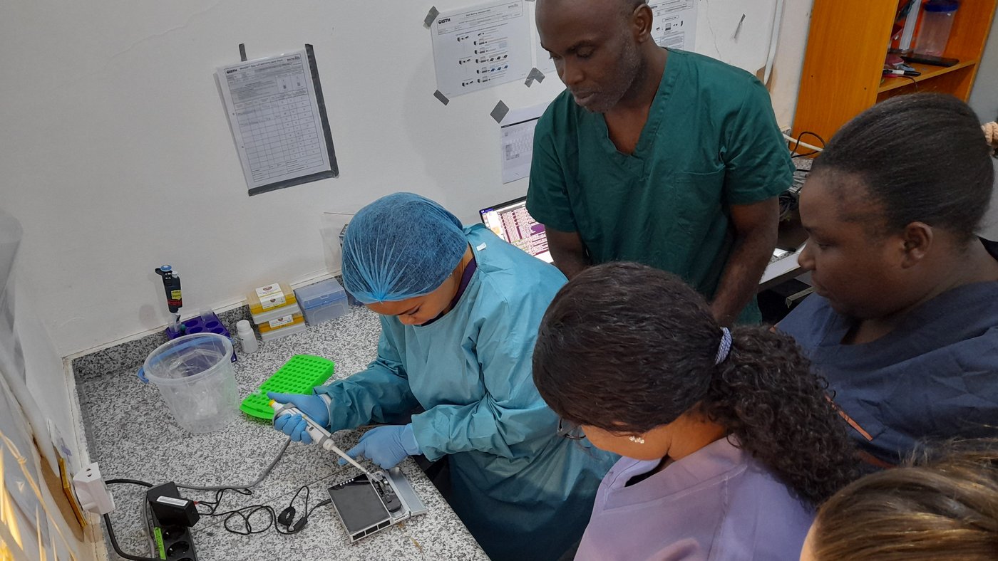 The photo shows a Nigerian lab team of four and their trainer at a lab bench. They wear blue and green lab coats and gloves. One of them is loading a sequencing device with a pipette while the others observe.