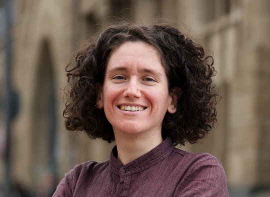 Dr. Giuditta Annibaldis Dr. Giuditta Annibaldis: eine lächelnde Forscherin mit dunklen Locken vor einer hellbraunen Häuserfassade. Sie trägt ein weinrotes Hemd und verschränkt die Arme vor dem Körper.