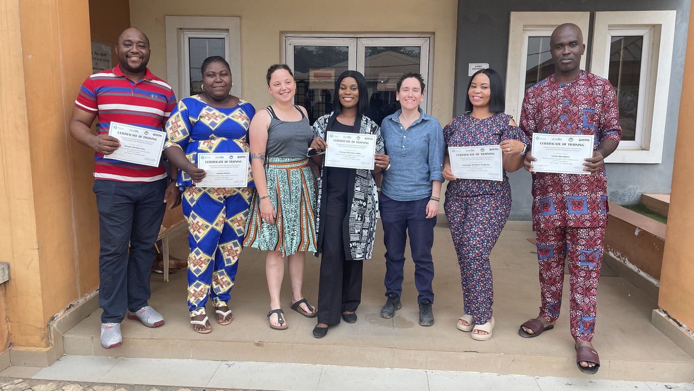 Five Nigerian laboratory staff and two BNITM trainers in colourful clothes are standing in the entrance of a building. They are proudly holding up their training certificates.