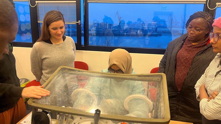 Arbeiten mit der Glove box Das Foto zeigt mehrere Wissenschaftler:innen um eine Glove box herum stehen. Eine von ihnen arbeitet gerade damit.