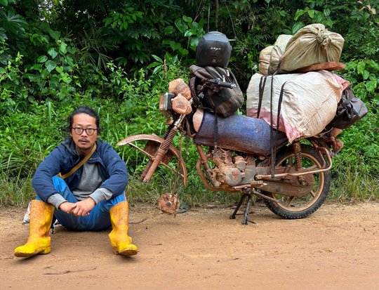 Sung Joon Park unterwegs in DRC Das Foto zeigt einen jungen Mann sitzend auf dem Boden neben einem vollbepackten Motorrad.
