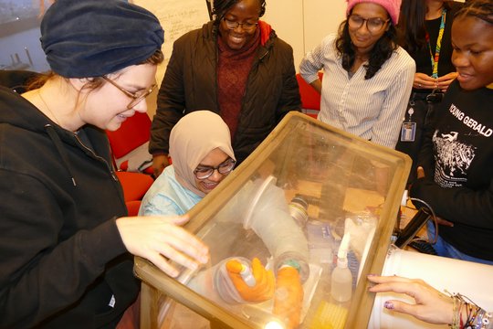 Glove box demonstration The foto shows 5 women standing around a glove box. One of them is training to handle materials in the glove box.