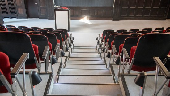 The photo shows the historic lecture theatre from behind with the red upholstered chairs in the foreground and the lectern in the foreground.