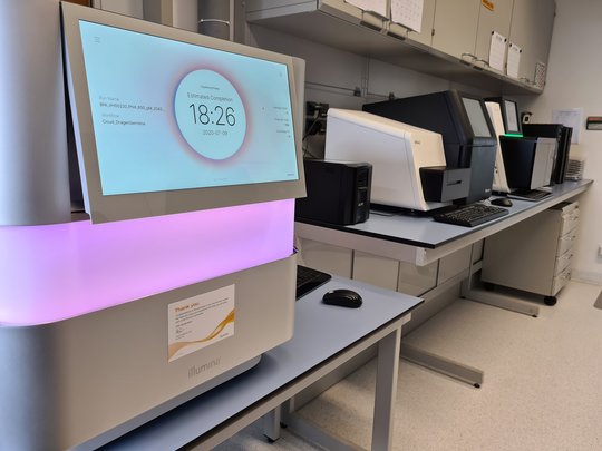 illumina der Next-Generation Sequencing (NGS) Einheit des BNITM Zu sehen ist die Next-Generation Sequencing (NGS) Einheit des BNITMs. Im Vordergrund steht ein großer weißer Kasten auf einem Tisch. in der Mitte verläuft ein breiter Leuchtstreifen, der in rosa-lila leuchtet. Oben sieht man einen großen Bildschirm, mit der Schrift estimated completion 18:26. Im Hintergrund stehen weitere Laborgeräte auf Tischen