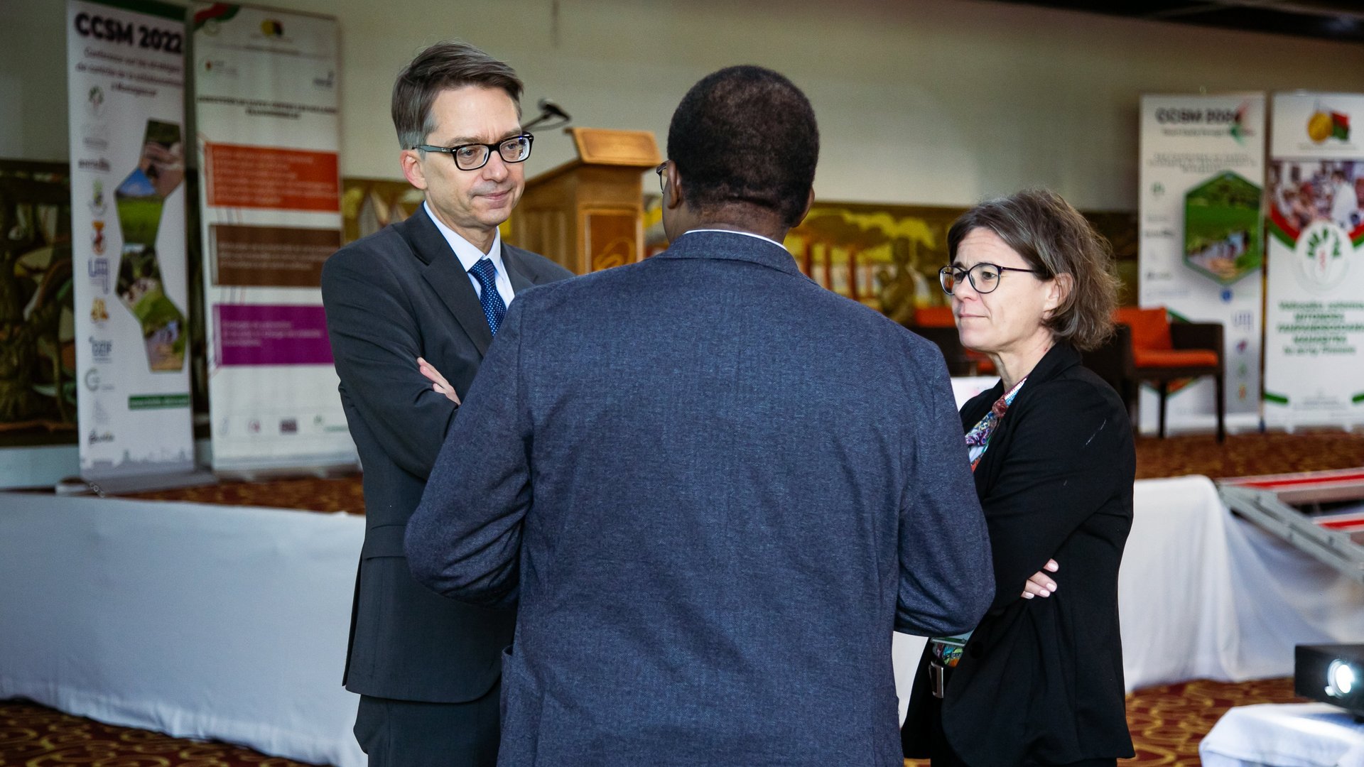 Three CCSM 2024 attendees engaged in a discussion near a stage, with event banners and a podium visible in the background, in a formal setting.