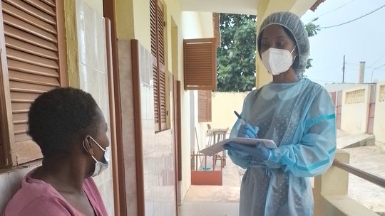 Patient interview Eine Person, die einen Laborkittel und eine Gesichtsmaske trägt, befragt eine Frau, die eine Gesichtsmaske trägt und auf einer Bank sitzt.