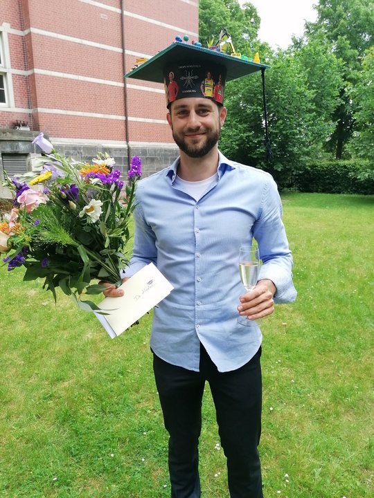 Stefan Hoenow Dissertation Stefan Hoenow kurz nach seiner Dissertation. Er trägt einen selbstgebastelten, bunten Doktorhut auf dem Kopf, in der rechten Hand einen großen Blumenstrauß und in der linken Hand ein Sektglas.