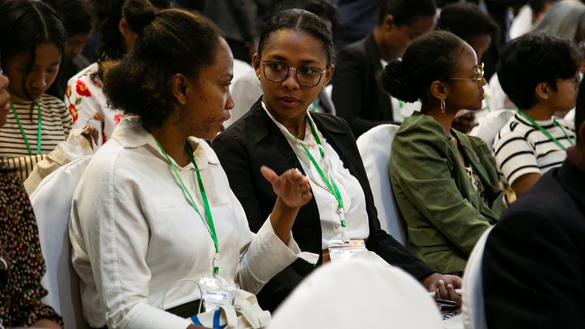 Two attendees seated in the CCSM 2024 conference discussing something, wearing event badges with green lanyards, surrounded by others in a formal setting.