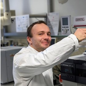 Konrad Langbecker, a student assistant with short blond hair, is shown wearing a lab coat, while operating the GC-MS and looking directly at the camera.