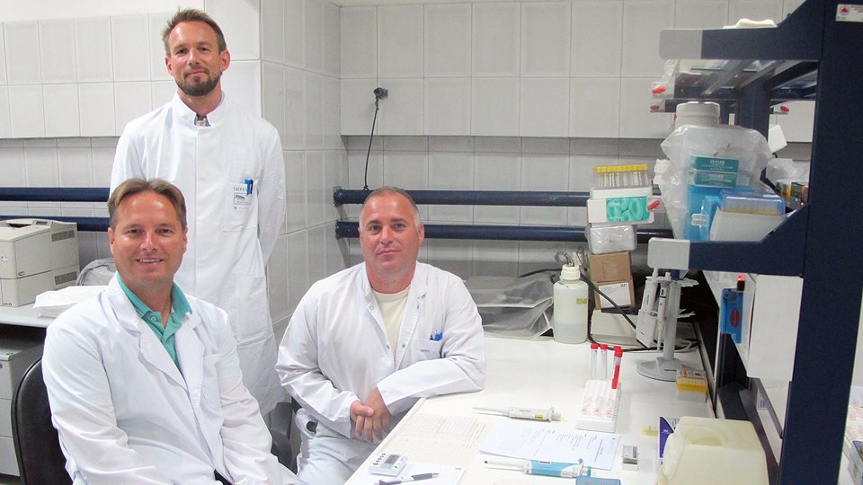 3 Forscher im Laborkittel sind im Labor zu sehen, zwei sitzen an einem Labortisch, auf dem unterschiedliche Laborutensilien und Notizen liegen. Ein dritter Forscher steht hinter den beiden sitzenden Forschern. Alle schauen in Richtung Kamera.