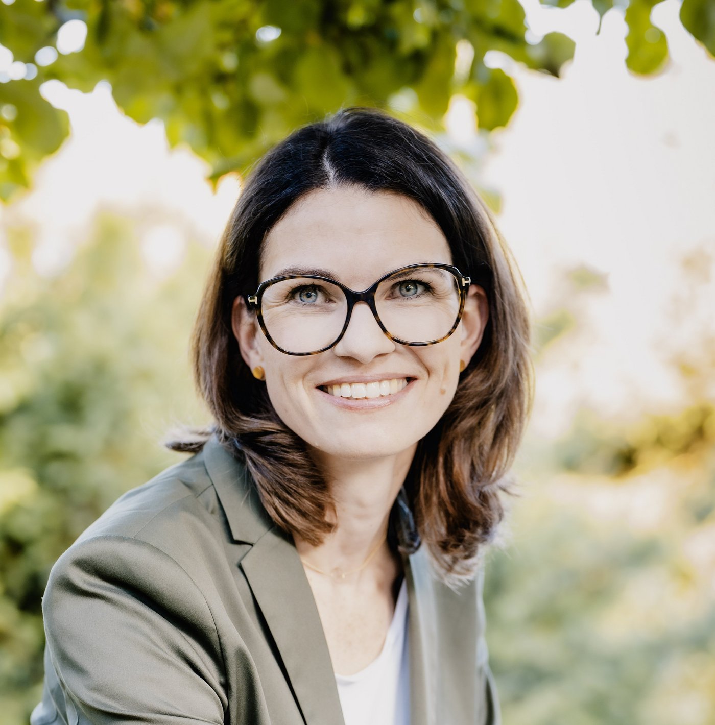 Prof. Dr. Cornelia Betsch: eine Forscherin mit braunen, schulterlangen Haaren, Brille und einem grauen Blazer über einem hellen Oberteil. Sie sitzt im Freien, im Hintergrund sind Blätter zu sehen.