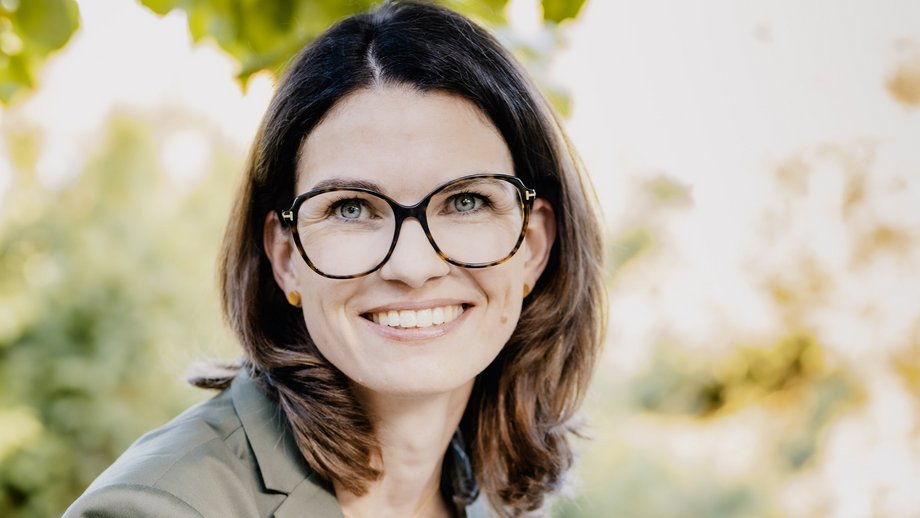 Prof. Dr. Cornelia Betsch: eine Forscherin mit braunen, schulterlangen Haaren, Brille und einem grauen Blazer über einem hellen Oberteil. Sie sitzt im Freien, im Hintergrund sind Blätter zu sehen.