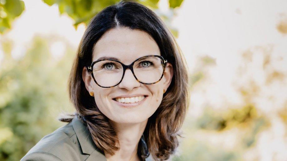 Prof. Dr. Cornelia Betsch: eine Forscherin mit braunen, schulterlangen Haaren, Brille und einem grauen Blazer über einem hellen Oberteil. Sie sitzt im Freien, im Hintergrund sind Blätter zu sehen.