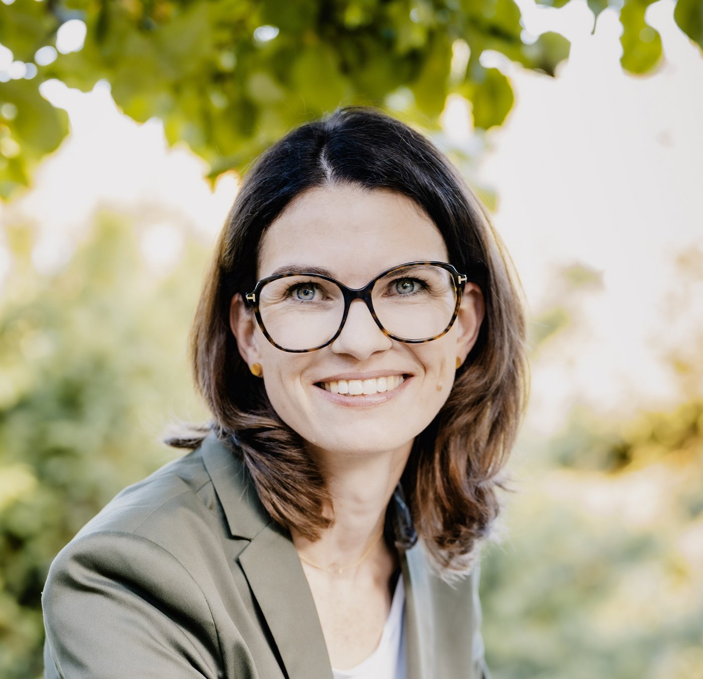 Prof. Dr. Cornelia Betsch: eine Forscherin mit braunen, schulterlangen Haaren, Brille und einem grauen Blazer über einem hellen Oberteil. Sie sitzt im Freien, im Hintergrund sind Blätter zu sehen.