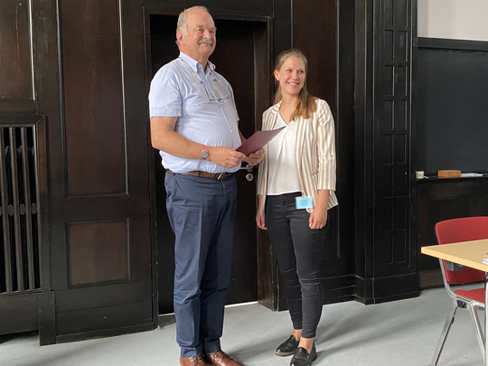 Marie Groneberg receive doctoral prizes from the BNITM Friends Association A board member of the Association of Friends of the Tropical Institute presents Marie Groneberg with a certificate at the award ceremony in the lecture theatre of the Bernhard Nocht Institute. In the background you can see a wall panelled in dark brown.