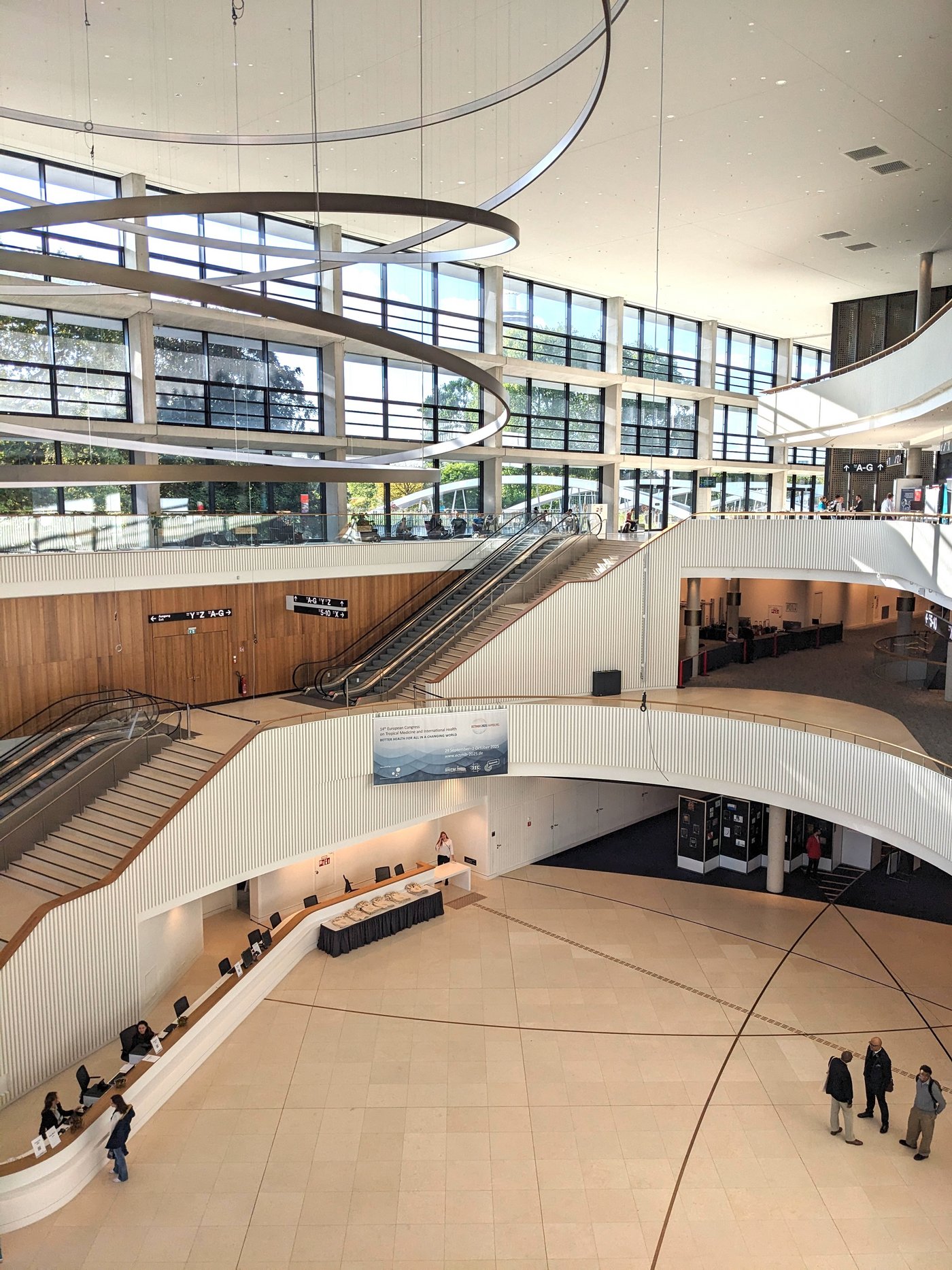 CCH - Congress Center Hamburg The picture shows the large reception hall of the CCH Hamburg. The staircase is very modern in beige and wood colors. A large lighting object made of steel circles hangs from the roof. The photo was taken from an upper floor.