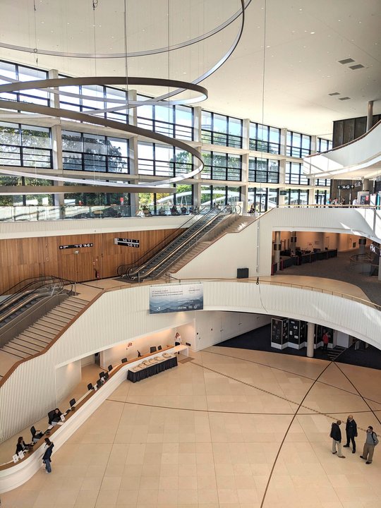 CCH - Congress Center Hamburg The picture shows the large reception hall of the CCH Hamburg. The staircase is very modern in beige and wood colors. A large lighting object made of steel circles hangs from the roof. The photo was taken from an upper floor.