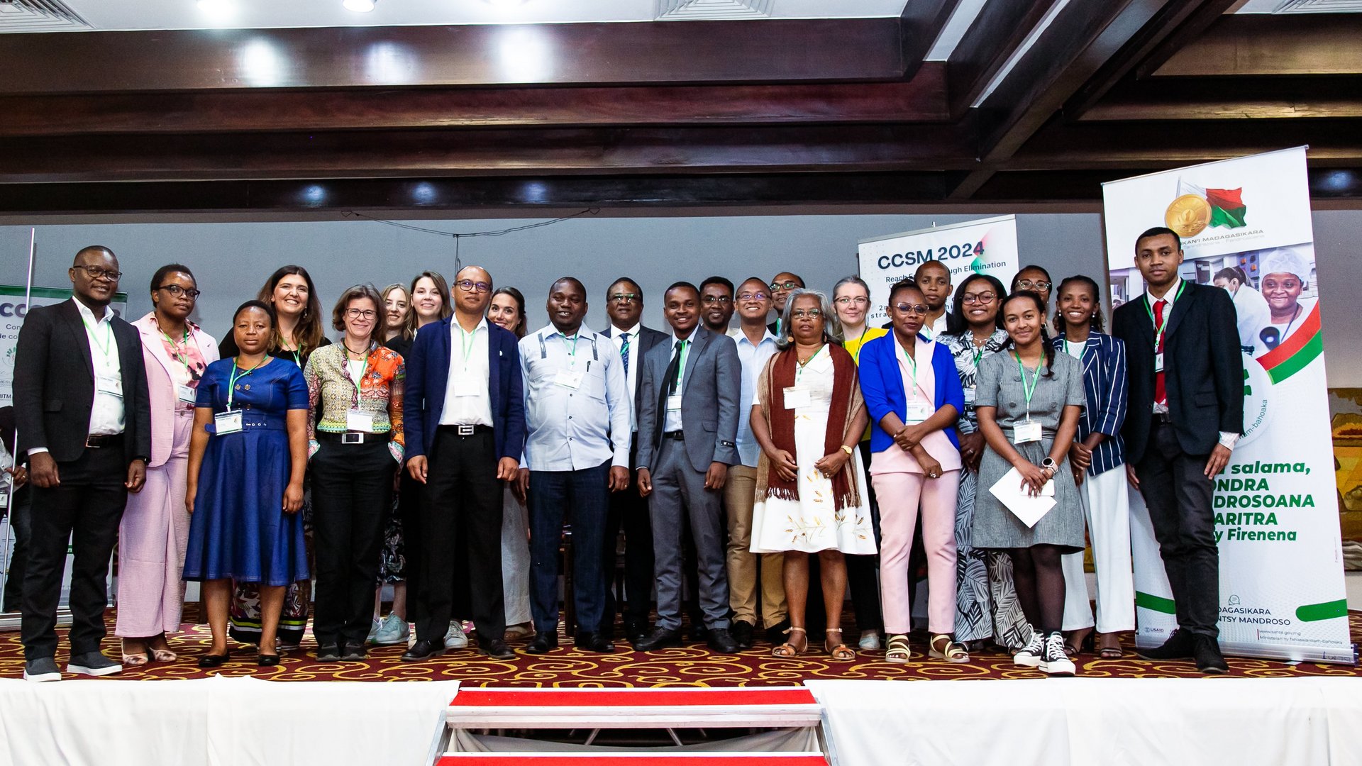 A large group of CCSM 2024 conference participants posing together on stage, with event banners in the background, wearing badges and formal attire.