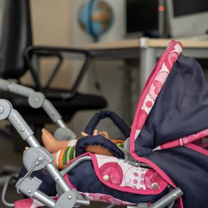 Das Familienzimmer Das Foto zeigt einen Spielzeug-Kinderwagen, im Hintergrund ein Schreibtisch-Arbeitsplatz.