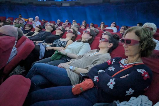 Space Tour 3D The picture shows visitors to the star show inside the planetarium. They are sitting back in comfortable armchairs and wearing 3D glasses. The lights have already been dimmed for the show.