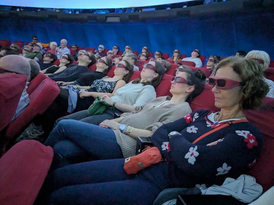 Space Tour 3D The picture shows visitors to the star show inside the planetarium. They are sitting back in comfortable armchairs and wearing 3D glasses. The lights have already been dimmed for the show.