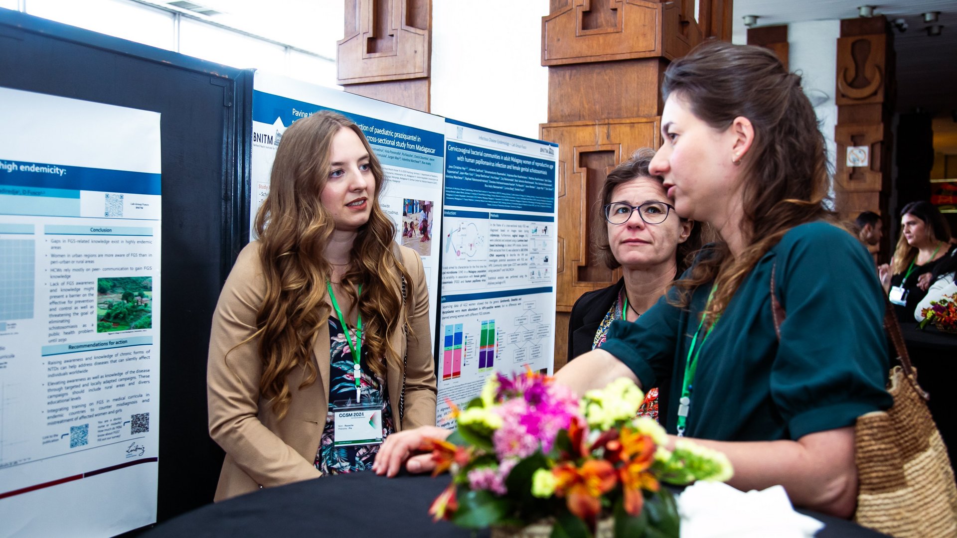 Three women engaged in a discussion in front of research posters at the CCSM 2024 conference, with a small floral centerpiece on a nearby table.