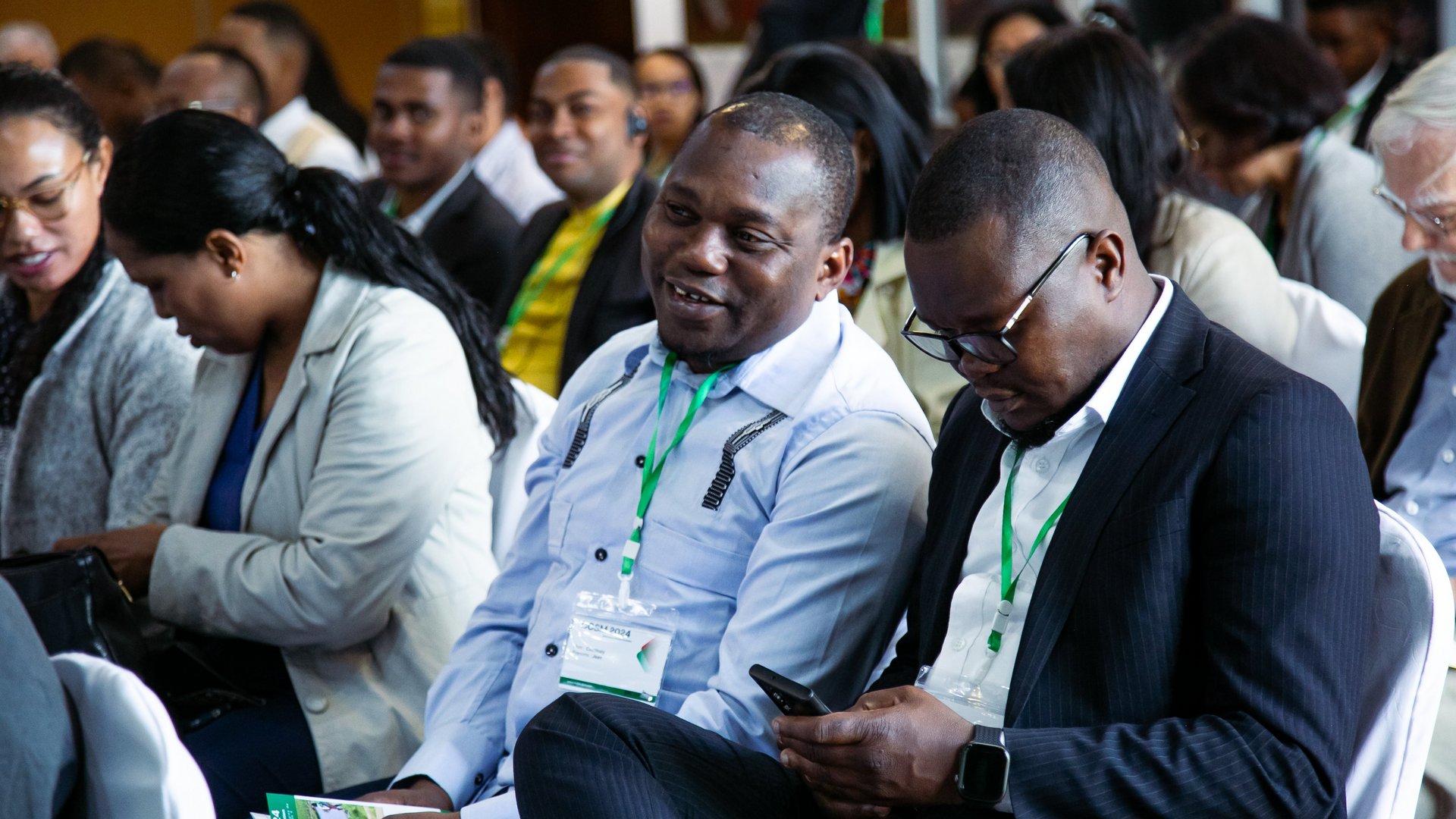 Two CCSM 2024 conference attendees seated, one smiling and the other focused on a phone, wearing green-lanyard badges, surrounded by others in a formal setting.