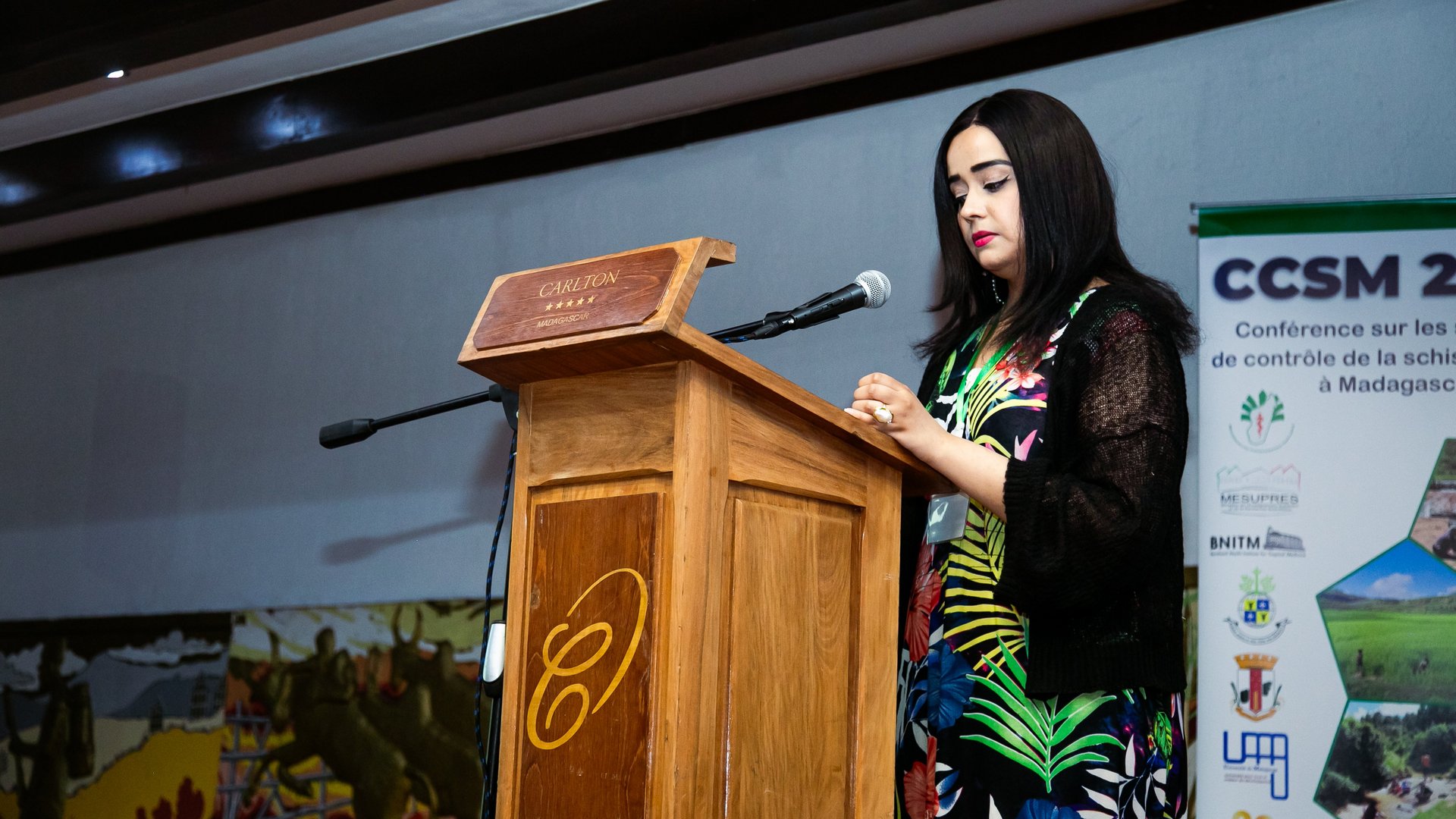 A woman presenting at the CCSM 2024 conference at a wooden podium, wearing a colorful dress, with a CCSM 2024 conference banner in the background.