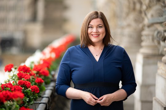 The Minister of Science and Deputy Mayor of the Free and Hanseatic City of Hamburg, Katharina Fegebank Katharina Fegebank: a politician on a balcony. On the left are red flowers, on the right the bright walls of Hamburg City Hall. Katarina Fegebank wears shoulder-length brown hair and a blue dress. In front of the center of her body, the fingertips of her two hands are touching.