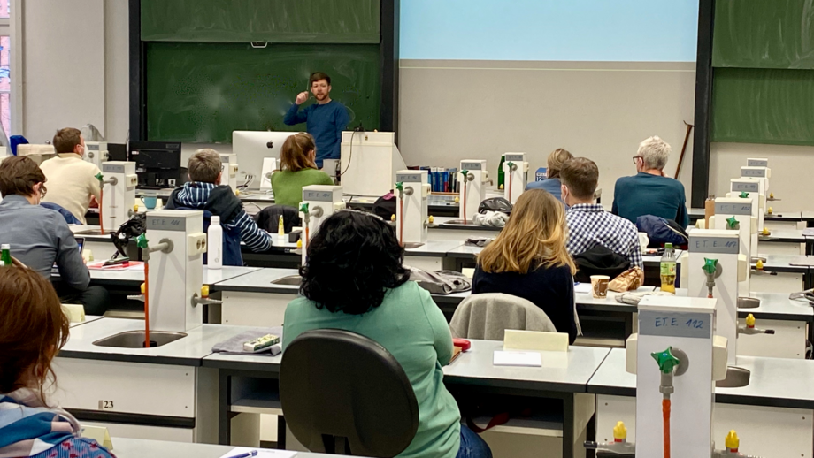 The photo shows the course room with course participants from behind. The green board can be seen in front, a lecturer is standing in front of it.